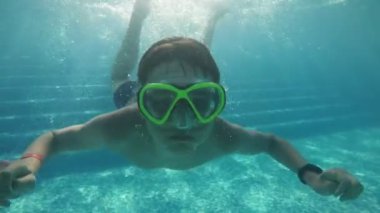 Mutlu çocuk su altında yüzüyor ve havuzda tek kollu amuda kalkıyor. Ağır çekimde neşeli bir esmer çocuk gözlüklü ve şortlu suyun altında yüzüyor ve tek kollu amuda kalkıyor. Yavaş çekimde havuzda ikiye bölünmüş bir şekilde.. 