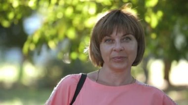 Yazın yeşil park alanında mutlu bir şekilde ayakta duran ve gülümseyen mutlu sarışın kadın pembe elbiseli neşeli sarışın kadın ve kısa saçlı kadın omuzunda süslü çantasıyla güneşli bir yaz parkında hayatının tadını çıkarıyordu. 