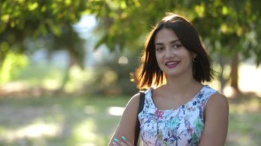 Güneşli bir günde yeşil parkta gülümseyen ve ellerini çaprazlayan neşeli esmer kız. Kolsuz açık renk elbiseli, gülümseyen ve elini yeşil park bölgesinde çaprazlayan saldırgan, romantik ve güzel esmer kız manzarası.
