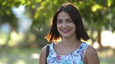 Yazın güneşli bir günde yeşil parkta dikilen ve gülümseyen neşeli esmer kız. Kolsuz, açık renk elbisesi olan güzel, romantik ve güzel esmer kız. Yazın yeşil bir park alanında ayakta, gülümsüyor ve akıllanıyor.