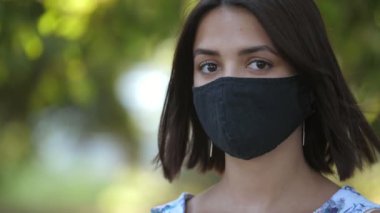 Güneşli bir günde yeşil parkta maske takan narin esmer kız kolsuz, açık renk elbiseli ve siyah maskeli güzel ve neşeli esmer bir kızın yaz mevsiminde yeşil park alanında ileriye bakması heyecan verici.