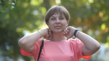 Gülümseyen sarışın kadın ayakta ve saçını düzeltiyor. Yavaş çekimde park ediyor. Güzel bir manzara. Şık, sarı bir kadın, pembe bir elbise ve kısa saç kesimi ile güneşli bir yaz gününde yeşil park alanında saçlarını tarıyor.