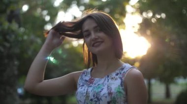Yazın günbatımında yeşil bir parkta akıllı akıllı akıllı bir kız. Açık renk kolsuz elbise giymiş güzel bir esmer kız ayakta duruyor, saçlarıyla oynuyor ve yazın günbatımında ağır çekimde yeşil bir parkta gülümsüyor..