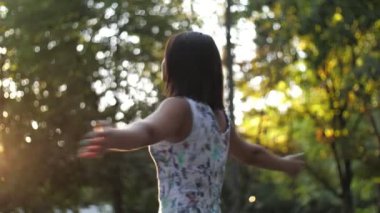 Romantik esmer kız yaz aylarında yavaş çekim parkında beyaz elbiseli, gülümseyen, dans eden ve yeşil bir parkta yavaşça gün batımını seyreden neşeli esmer kız manzarasına bakıyor.