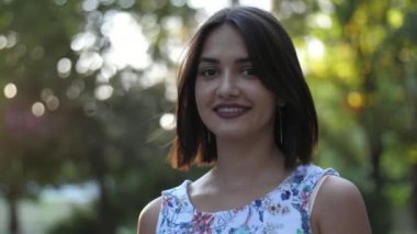 Yazın günbatımında parkta ayakta duran ve hayatın tadını çıkaran mutlu bir kız. Açık beyaz elbiseli neşeli esmer bir kız, gülümseyen, ayakta duran ve hayatının tadını çıkaran. Yavaş çekimde parlayan yeşil bir parkta.