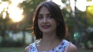Güzel bir kız, yaz ayının altın gün batımında parkta ayakta ve gülümsüyor. Yavaş çekimde, parlak bir esmer kız, yaz elbisesi içinde gülümsüyor ve hayatının tadını çıkarıyor..