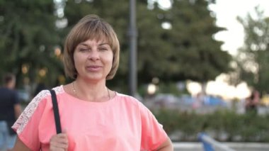 Başarılı bir kadın, gül rengi elbisesiyle yazın yavaş çekimde rıhtımda gülümseyen ve ayakta duran mutlu, pembe elbiseli, siyah çantalı bir kadının güneşli bir yaz gününde Dinyeper Nehri rıhtımında gülümseyen ve ayakta duran muhteşem manzarası. 