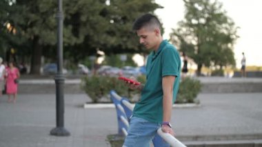 Nehir kıyısında duran ve yazın ağır çekimde internette gezinen genç adam Dnipro nehri rıhtımında tırabzanlara yaslanan ve ağır çekimde telefonuyla internette sörf yapan kısa saçlı, yeşil tişörtlü genç adamın etkileyici yan görüntüsü.. 