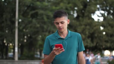 Yaz mevsiminde yeşil Dnipro nehri rıhtımında elinde ağır çekimde kırmızı akıllı telefonla dolaşan kısa saçlı, kısa saçlı, sporcu görünümlü, neşeli genç adam, nehir kenarında yürüyor ve internette sörf yapıyor. İnternette geziniyor.