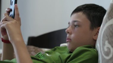 Rahat çocuk bir dairede akıllı telefonunda yalan söylüyor ve video oyunları oynuyor. Kısa saçlı, yeşil tişörtlü ve dinlenen esmer bir çocuğun modern bir apartmandaki akıllı telefonunda uzanmış video oyunları oynaması.. 