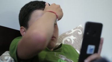 Akıllı çocuk kanepede uzanıyor ve evinde akıllı telefonuyla internette geziniyor. Tişört giymiş, şort giymiş, kısa saçlı, akıllı bir esmer çocuğun evdeki telefonuyla interneti tırmalıyor ve internette geziniyor.. 