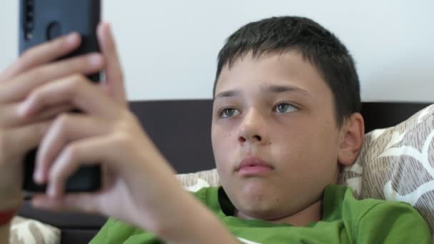 不满的男孩躺在公寓里 用他的现代照片玩电子游戏 这是个不同寻常的景象 一个有着短发的轻松的黑发男孩 穿着一件绿色T恤躺在沙发上 在一个通风的公寓里用智能手机玩电子游戏 — 图库视频影像