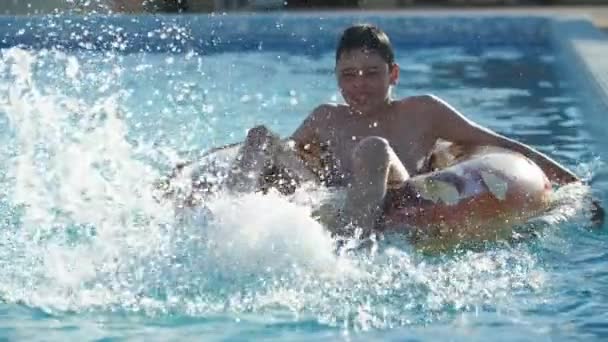 楽しい男はゆっくりとした動きで光沢のあるスイミングプールで足で水を散らします水の底に座っているブルネットの少年の素晴らしい景色 笑顔と遅い動きで日当たりの良い日にプールで水車の動きを作ります — ストック動画