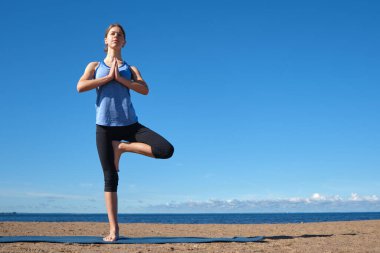 Genç, sıska bir kız, güneşli bir sabahta plajda yoga yapıyor, güneş selamı veriyor, spor yapmadan önce esniyor.
