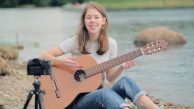 Genç bir kadın blogcu elinde gitarla gölün kenarında oturuyor.