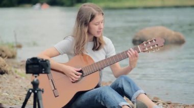 Genç bir kadın blogcu elinde gitarla gölün kenarında oturuyor.