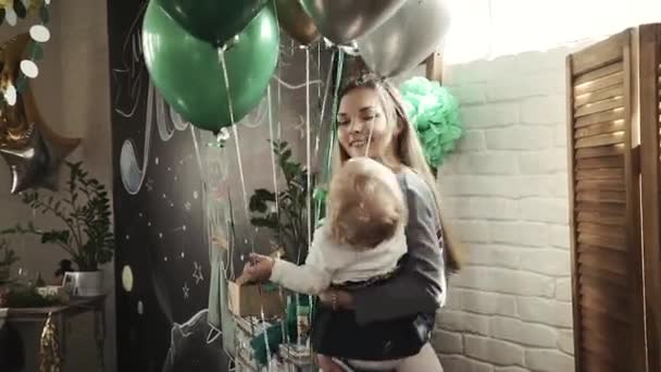 Junge Mutter hält eine einjährige Tochter, die zum Geburtstag mit Luftballons spielt — Stockvideo