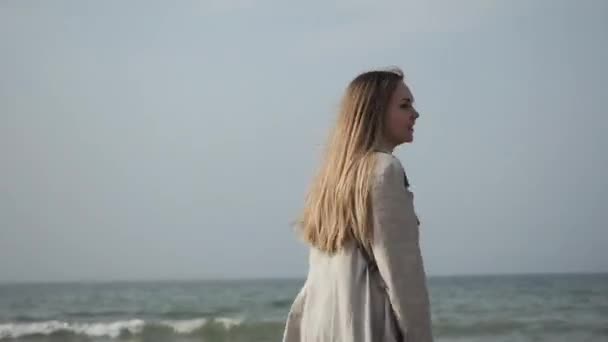 Νεαρή γυναίκα με αδιάβροχο που χορεύει στην παραλία της θάλασσας, περπατώντας κατά μήκος της ακτής — Αρχείο Βίντεο
