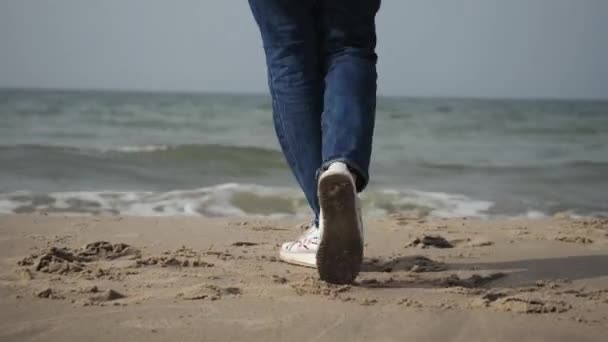 Primer plano de una joven con un abrigo beige y vaqueros azules bailando en la playa del mar, caminando por la costa — Vídeos de Stock