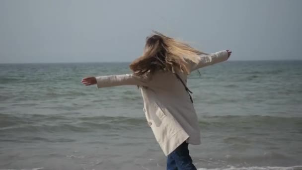 Молодая женщина в дождевике танцует на берегу моря, гуляет по побережью — стоковое видео