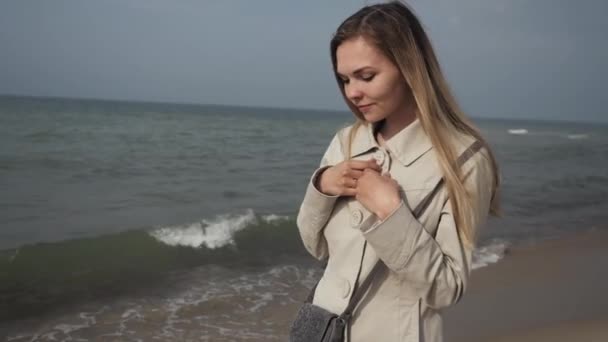 Молодая счастливая женщина с длинными волосами в бежевом пальто крупным планом танцует на морском пляже — стоковое видео