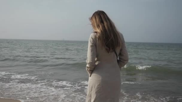 Junge glückliche Frau mit langen Haaren im beigen Mantel in Großaufnahme, die am Strand tanzt — Stockvideo