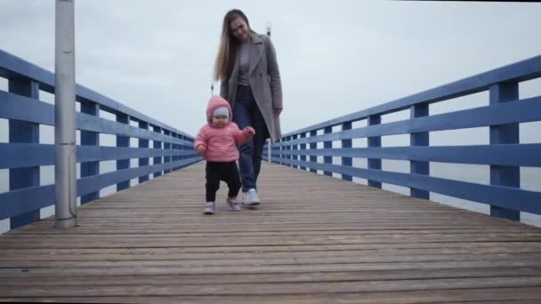 Молодая счастливая женщина мама идет с маленькой девочкой, целует свою дочь вдоль набережной моря первые шаги — стоковое видео
