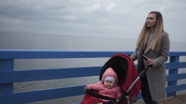 Genç anne, deniz kenarında bir kadın, içinde küçük bir kızın kızı olan bir bebek arabasıyla yürüyor.