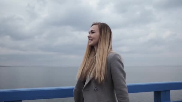 Giovane donna felice bionda con i capelli lunghi in un cappotto grigio e jeans passeggiate lungo il lungomare — Video Stock