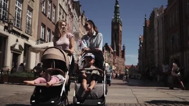 Jovens mães felizes mulheres com carrinhos de bebê caminham pela rua falando caminhada — Vídeo de Stock