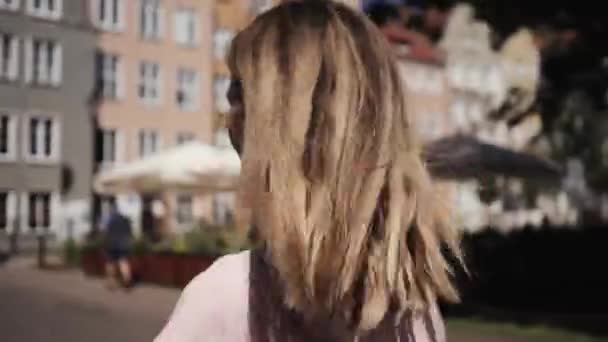 Νεαρή ξανθιά γυναίκα με μακριά μαλλιά βόλτες γυρίζει γύρω, κουνάει το κεφάλι της με τα μαλλιά, πίσω όψη — Αρχείο Βίντεο