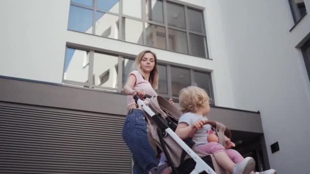 Young mother walks with a little girl with her daughter in a baby carriage near the house — Stock Video