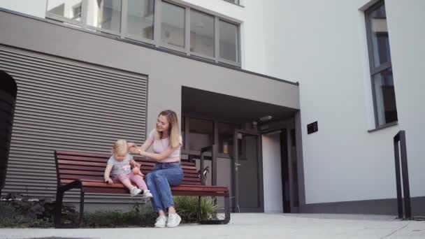 Jovem mãe caminha com sua filha menina em torno da casa sentada em um banco falando — Vídeo de Stock