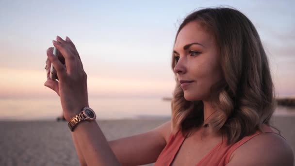 Όμορφη νεαρή γυναίκα παίρνει φωτογραφίες σε ένα smartphone σε ένα φόρεμα που ποζάρει βόλτες κατά μήκος της ακτής χαμογελώντας στο ηλιοβασίλεμα κοντά στη θάλασσα — Αρχείο Βίντεο