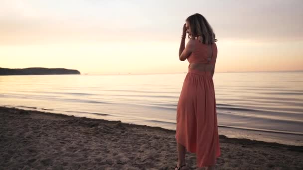 Bela jovem mulher em um vestido posando ao longo da costa sorrindo ao pôr do sol perto do mar — Vídeo de Stock