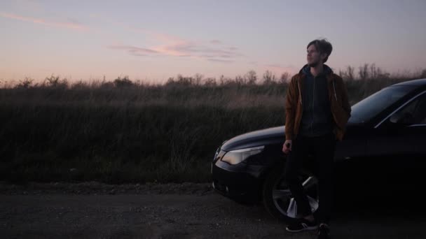 Brooding olhar para a distância que o jovem olha à noite ao lado de um carro — Vídeo de Stock