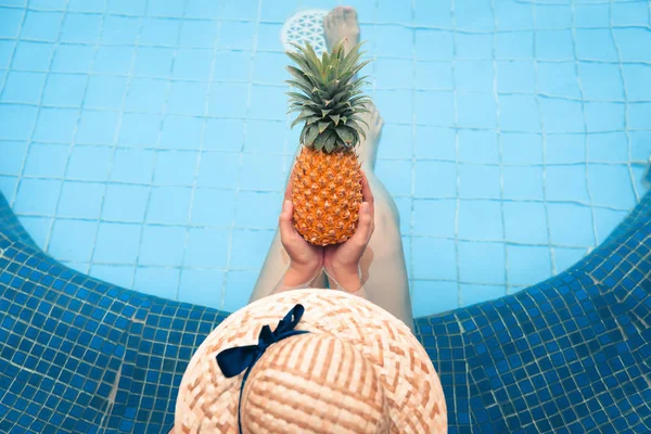 Portrait of beautiful woman relaxing in swimming pool while holding a pineapple, Recreation sport outdoor and summer vacation., Pretty woman in swimsuit with her hat relaxing sunbathing beside a pool.