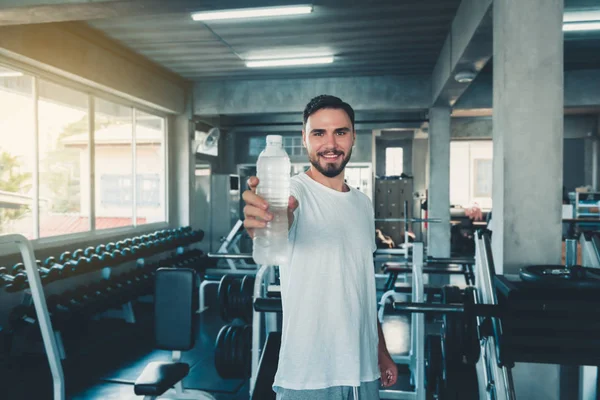 Portrait of sporty male holding bottle of water to giving someone in front of camera on exercise equipment background., Smiling sports man working out in gym.,Sport club and healthy concept.