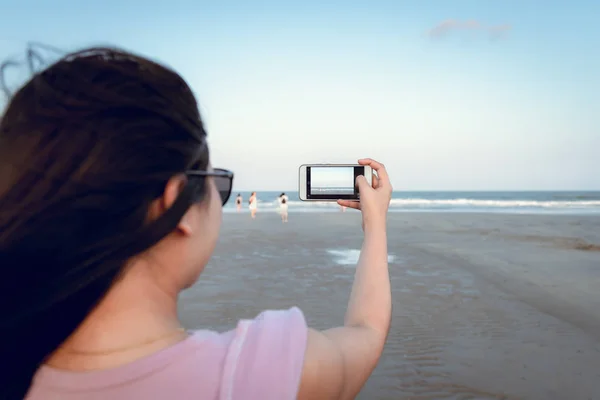 Traveling and Technology Concept, Woman is Taking Photos at The Beach by Her Smartphone in Summer Vacation. Asian Tourist is Relaxing With Her Cell Phone on The Beach in Holiday time.