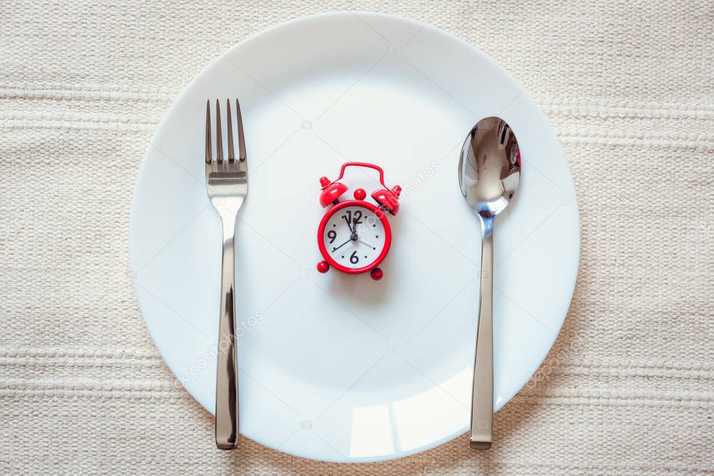 Healthy Eat Meal Time and Reminder Breakfast Concept, Food Timing Cycles for Eating, Red Alarm Clock With Stainless Steel Spoon and Fork on Ceramic Dish. Empty Plate and Household Object on a Table
