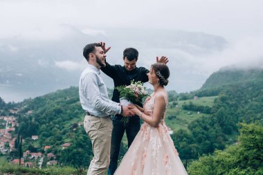 Yeni evliler doğada yürüyüş. Damat ve gelin dağlar ve deniz arka planı. Genç rahip kutsar.