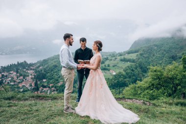 Yeni evliler doğada yürüyüş. Damat ve gelin dağlar ve deniz arka planı. Genç rahip kutsar.