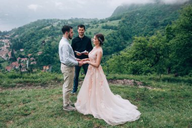 Yeni evliler doğada yürüyüş. Damat ve gelin dağlar ve deniz arka planı. Genç rahip kutsar.