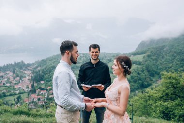 Yeni evliler doğada yürüyüş. Damat ve gelin dağlar ve deniz arka planı. Genç rahip kutsar.