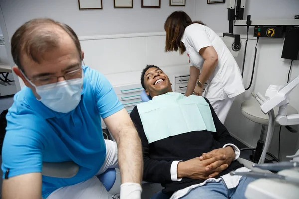Bir dişçi Kliniği ve yordam için hemşire ile hazırlama sandalyede oturan Afrikalı-Amerikalı adam gülüyor.