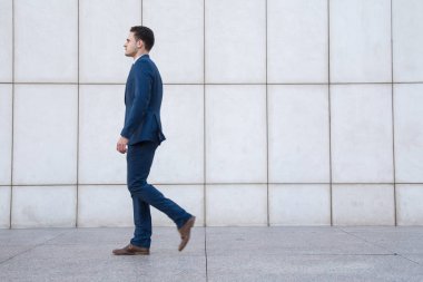 Takım elbise ve ufuk yüz sokakta bir beyaz duvar arka plan üzerinde yürüme ile genç iş adamı.