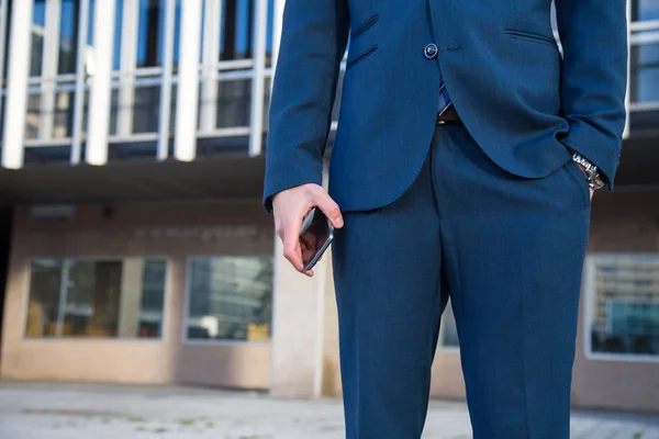 Faceless shot of man in trendy suit holding phone while standing with hand in pocket on street.