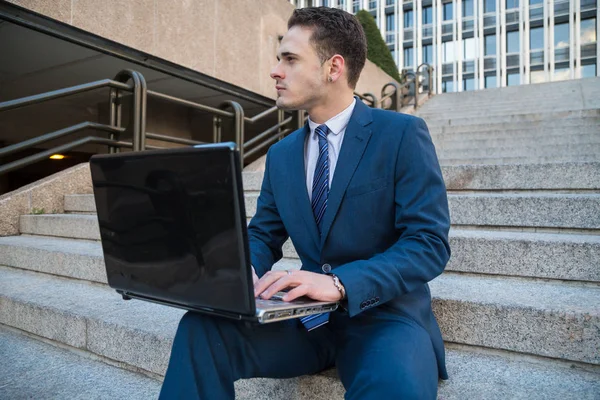 Dreaming man in elegant suit posing on stairs with laptop on knees looking away.