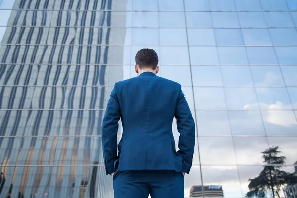 Back view of businessman in classy suit standing with hands in pockets on background of glass building.