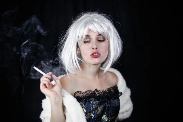 Attractive young woman in costume smoking cigar and looking at camera while standing on black background